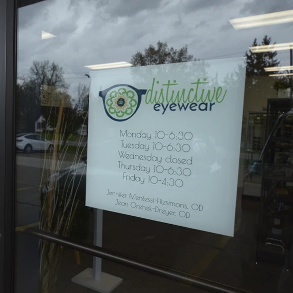Storefront window displaying Distinctive Eyewear’s business hours and branding.